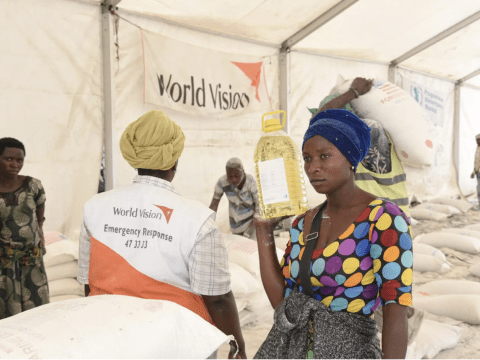 A woman holds a bottle of cooking oil while standing near sacks of food at a distribution point